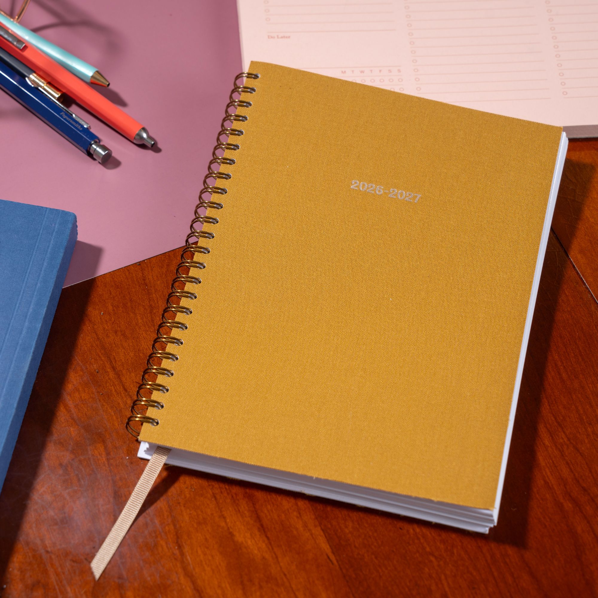 Yellow spiral-bound planner on a wooden surface with pens and another book in the background.