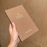 Brown journal titled 'The Five Minute Journal' held by a hand against a wooden background