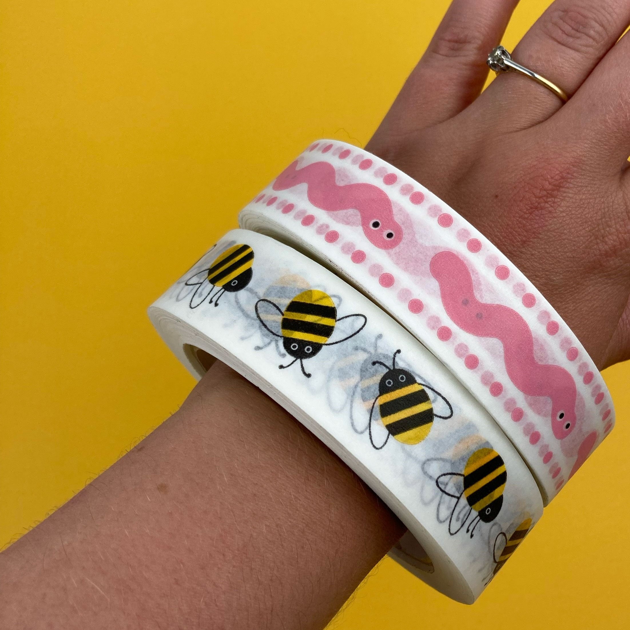 Hand holding two rolls of tape with bee and pink worm designs on a yellow background