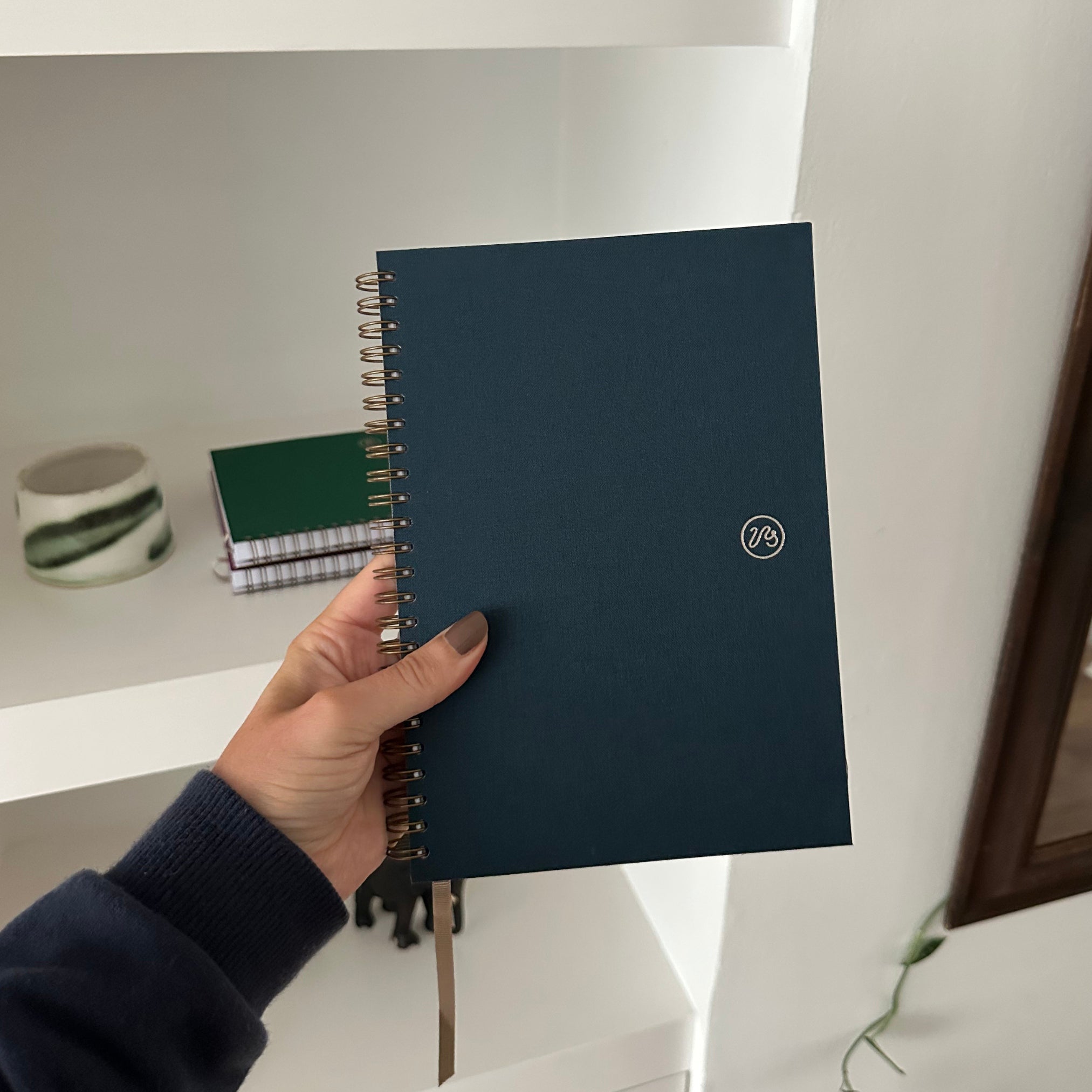 Hand holding a dark blue notebook with a logo on a blurred indoor background