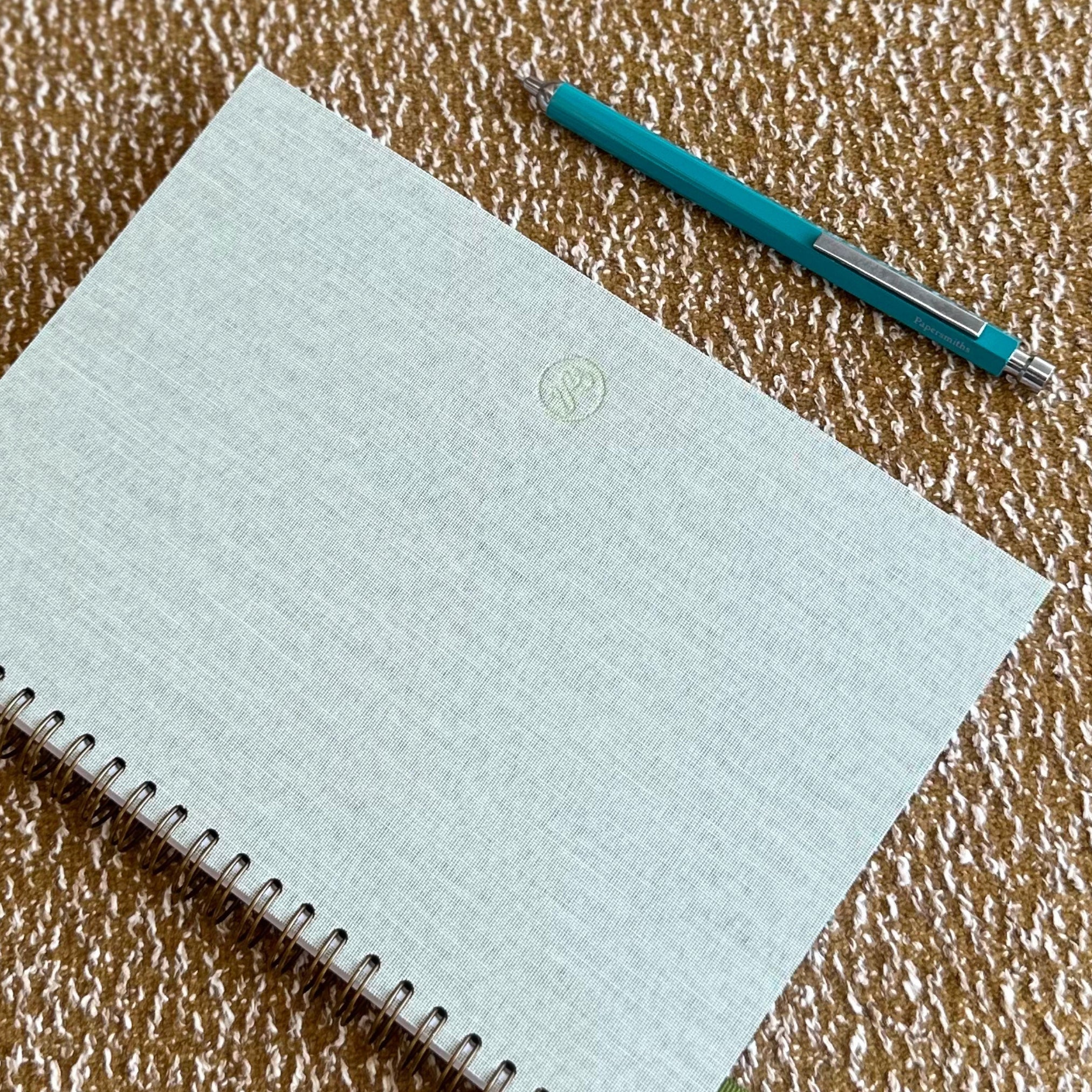 Light blue notebook with a green logo on a textured brown surface, accompanied by a blue pen.