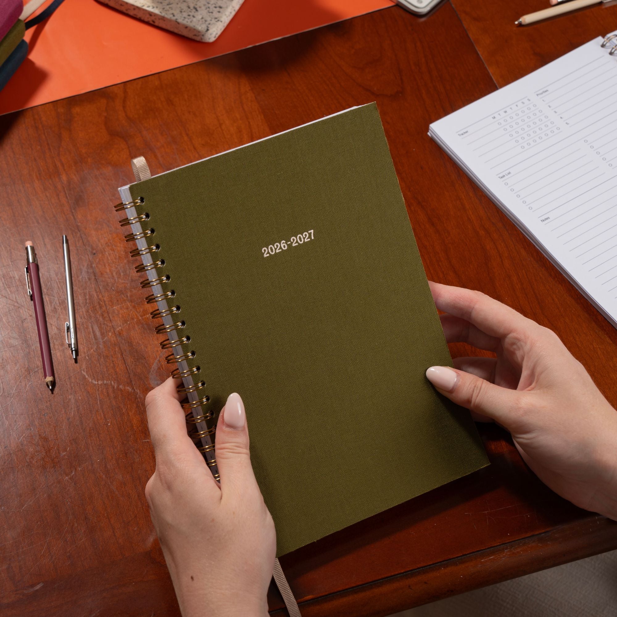Person holding a green spiral-bound planner on a wooden desk with stationery items.