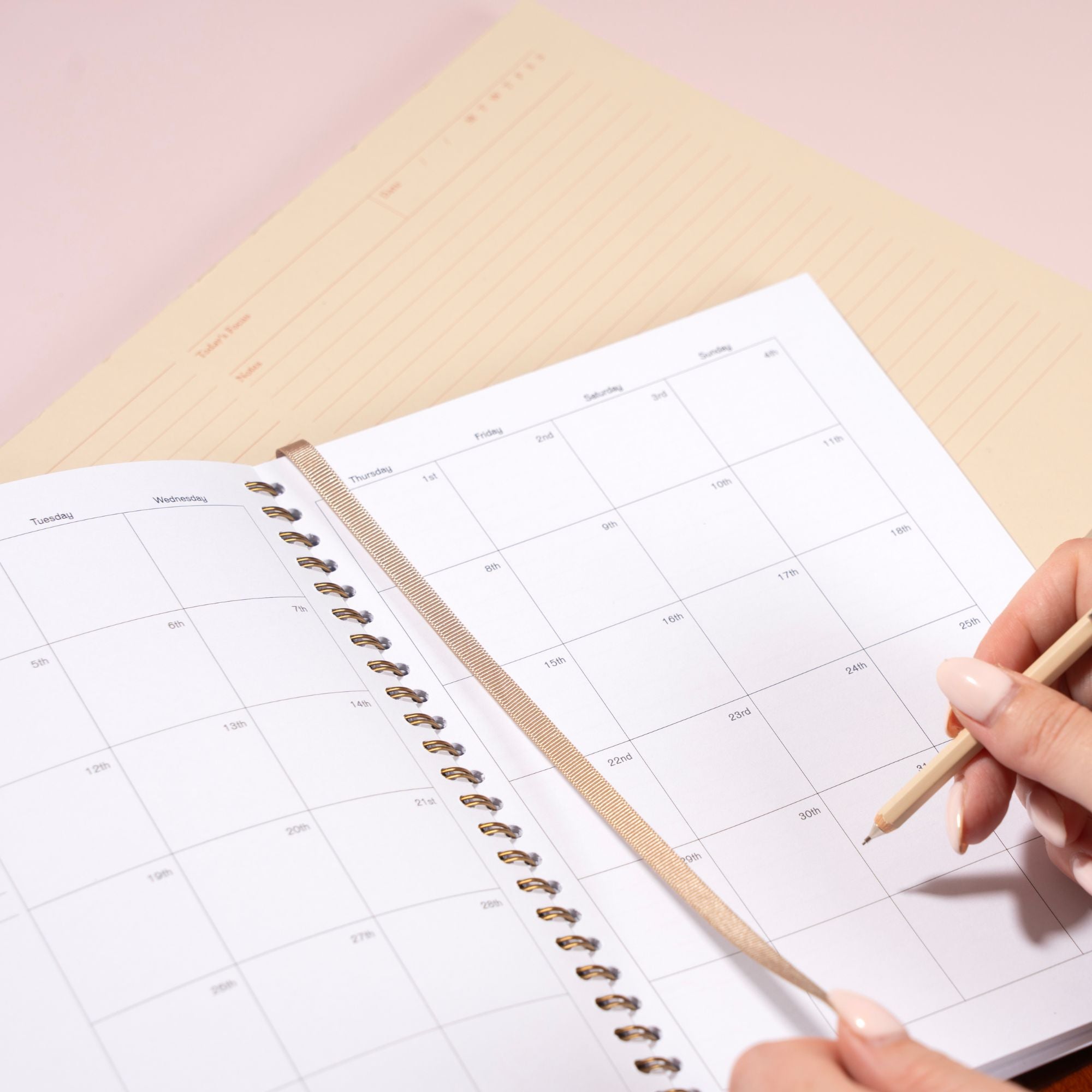 Person holding a pencil over a planner with a grid layout on a light pink surface
