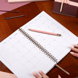 Spiral-bound planner on a wooden table with a pink pen and hands holding it.