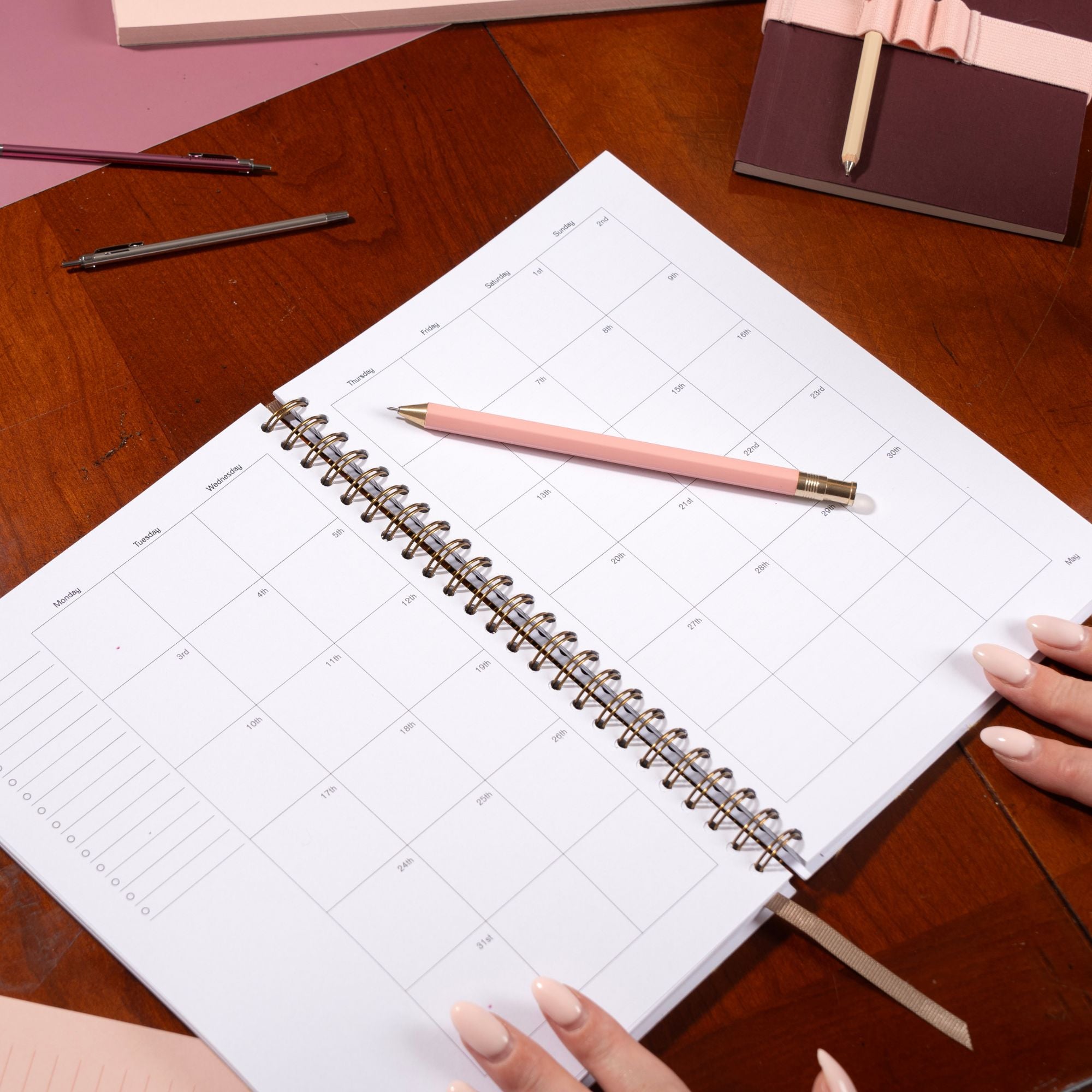 Spiral-bound planner on a wooden table with a pink pen and hands holding it.