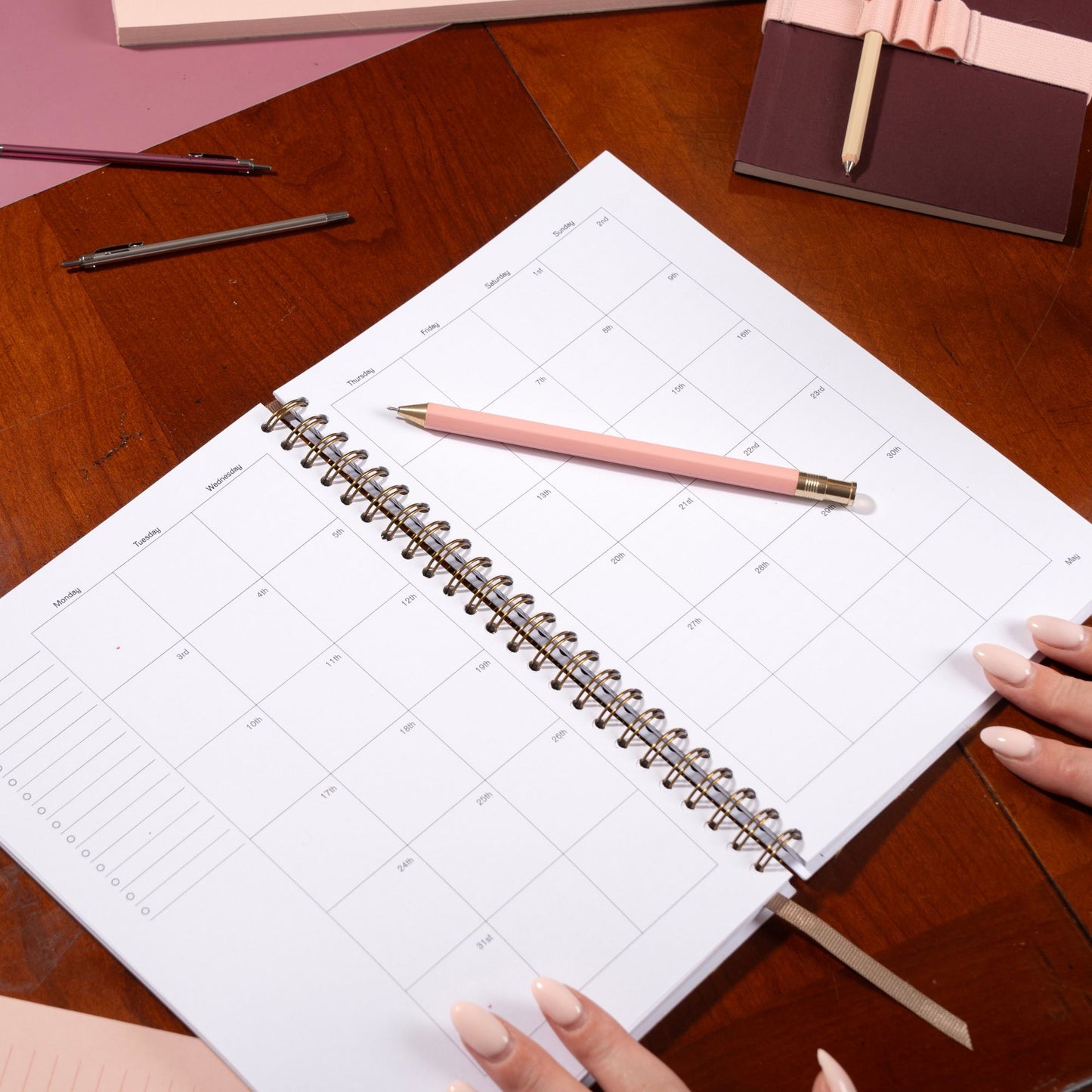 Spiral-bound planner on a wooden table with a pink pen and hands holding it.