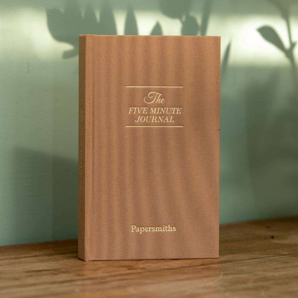 Brown journal titled 'The Five Minute Journal' by Papersmiths on a wooden surface with a blurred green background