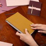 Brown notebook on a wooden table with a hand holding it, surrounded by other notebooks and pens.