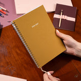 Ochre planner with 2026-2027 text held by a hand on a wooden surface with other planners and pens.