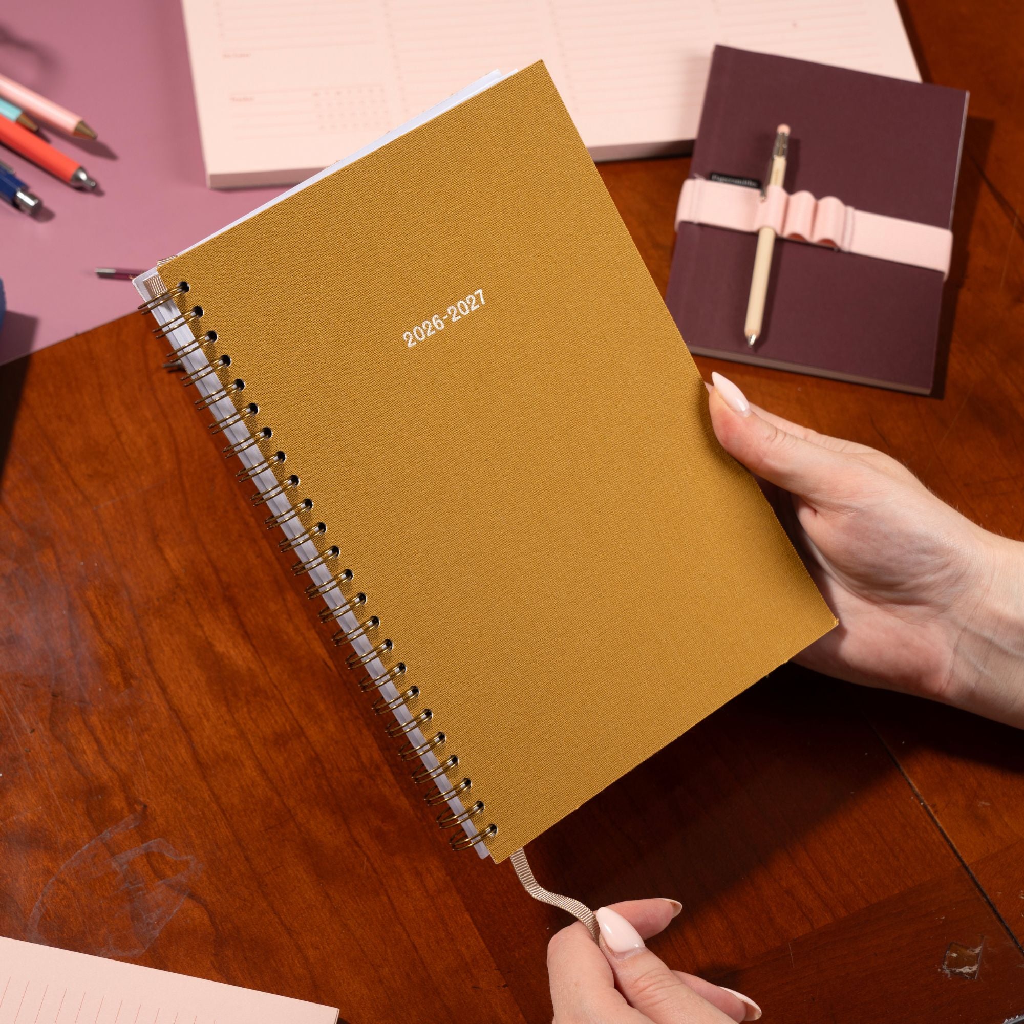 Ochre planner with 2026-2027 text held by a hand on a wooden surface with other planners and pens.