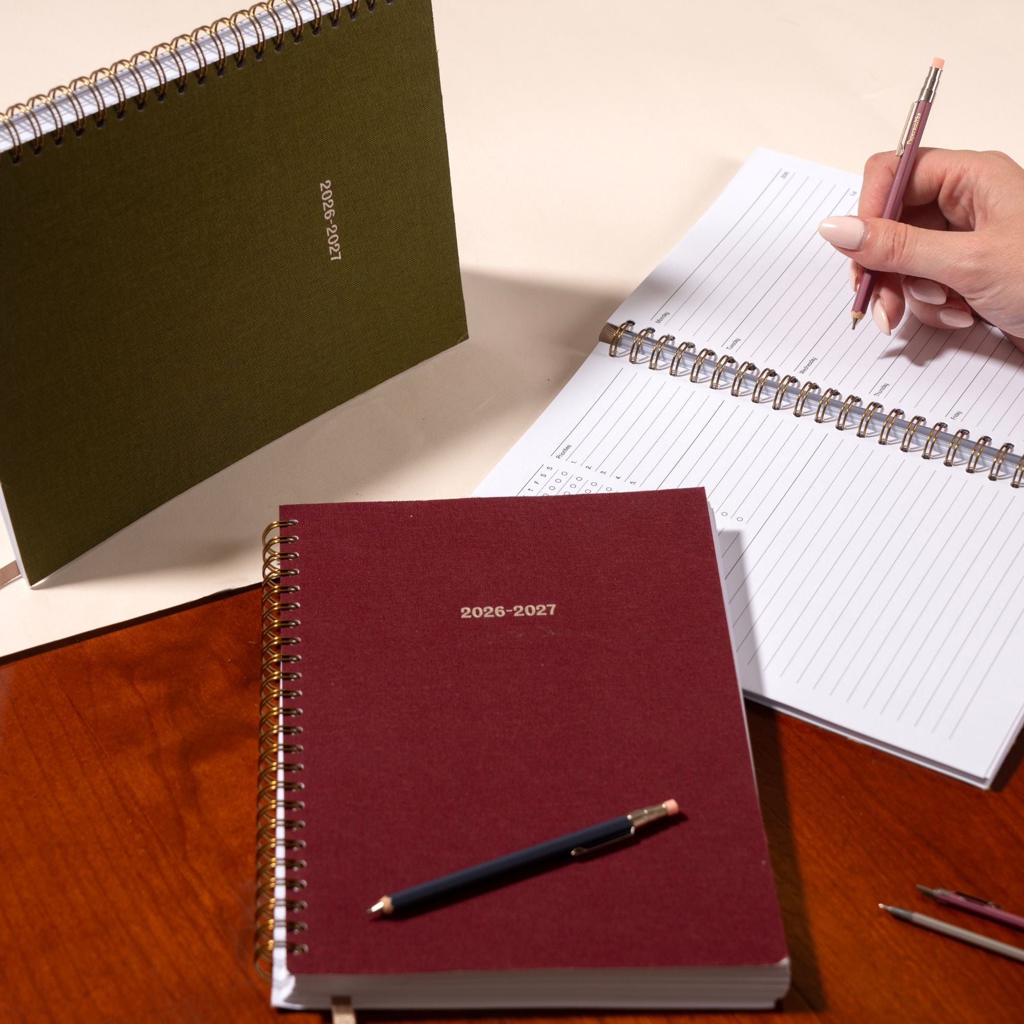 Maroon planner with 2026-2027 text, pen, and open planner on a wooden surface.