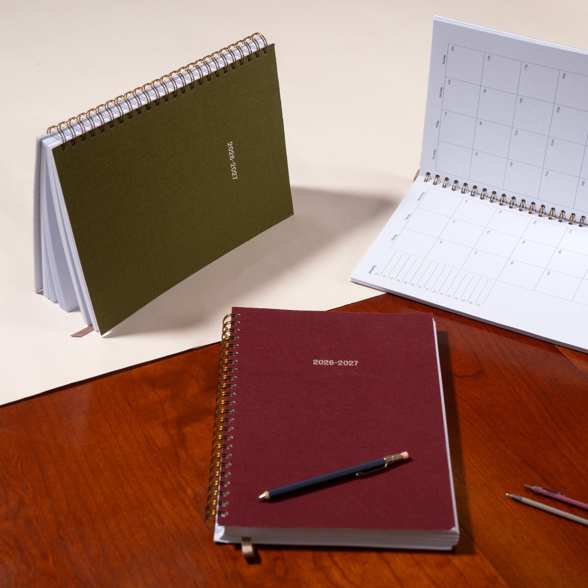 Three spiral-bound planners on a wooden surface with a pen.