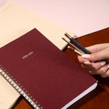 Maroon planner with '2026-2027' text, held by a hand with pens on a wooden surface.