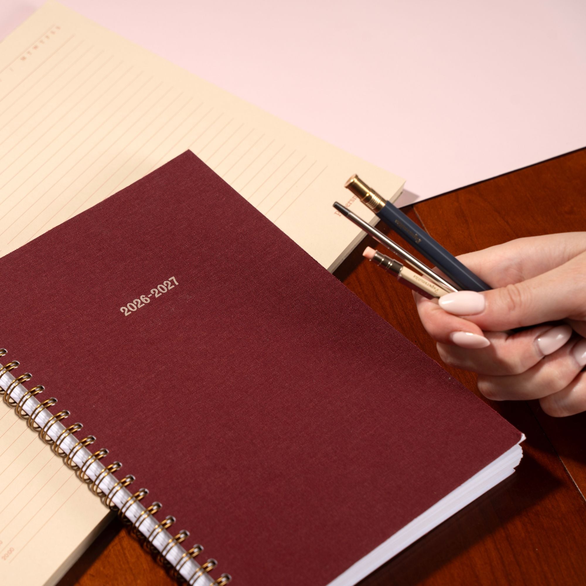 Maroon planner with '2026-2027' text, held by a hand with pens on a wooden surface.