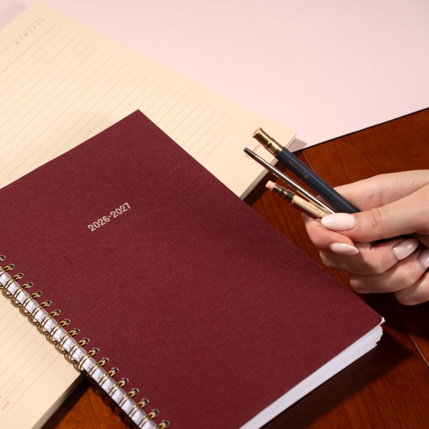 Maroon planner with '2026-2027' text, held by a hand with pens on a wooden surface.