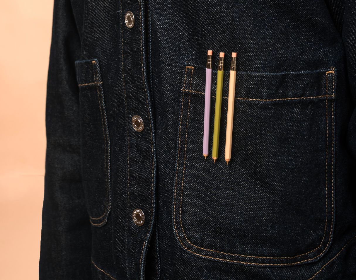 Three colorful pencils in a dark denim jacket pocket against a peach background