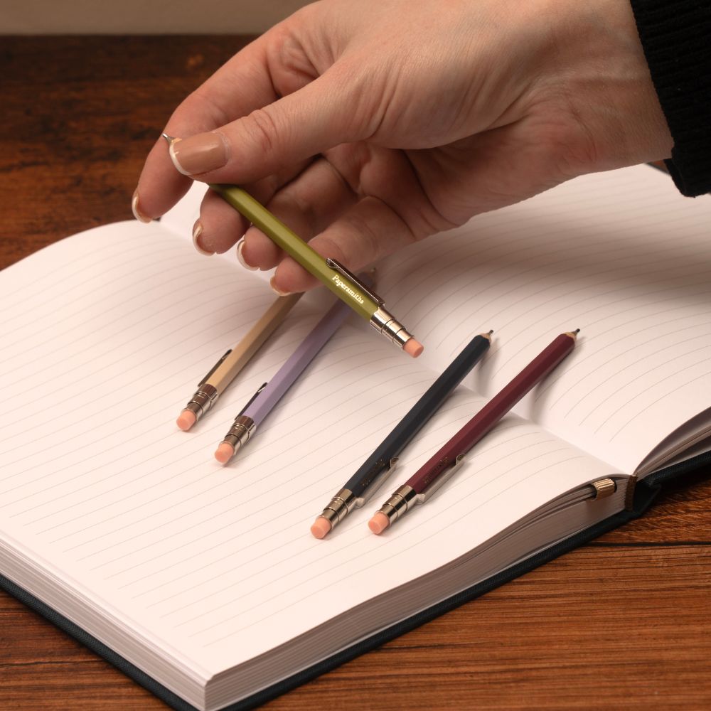 Hand holding a pencil over an open book with other pencils on the page.