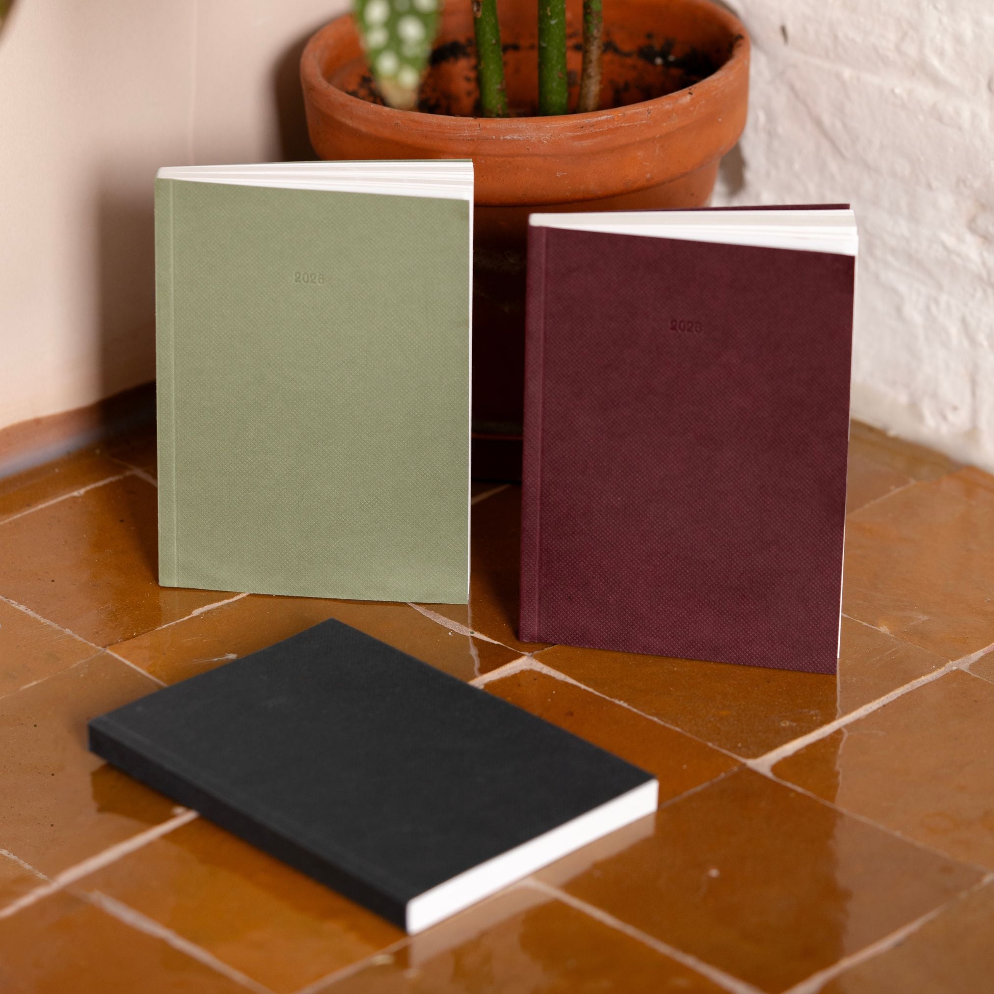 Three notebooks in green, maroon, and black on a tiled floor with a potted plant in the background.