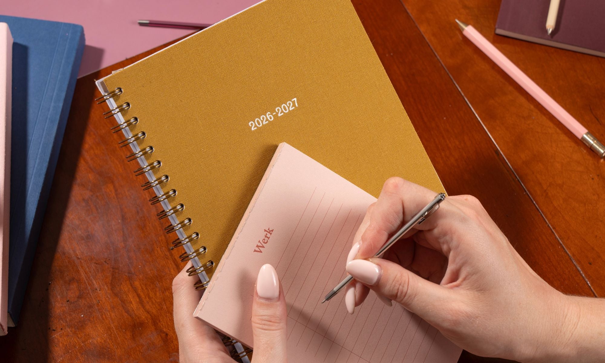 Person writing in a pink notebook with a 2026-2027 planner on a wooden table.