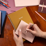 Person writing in a pink notebook with a 2026-2027 planner on a wooden table.